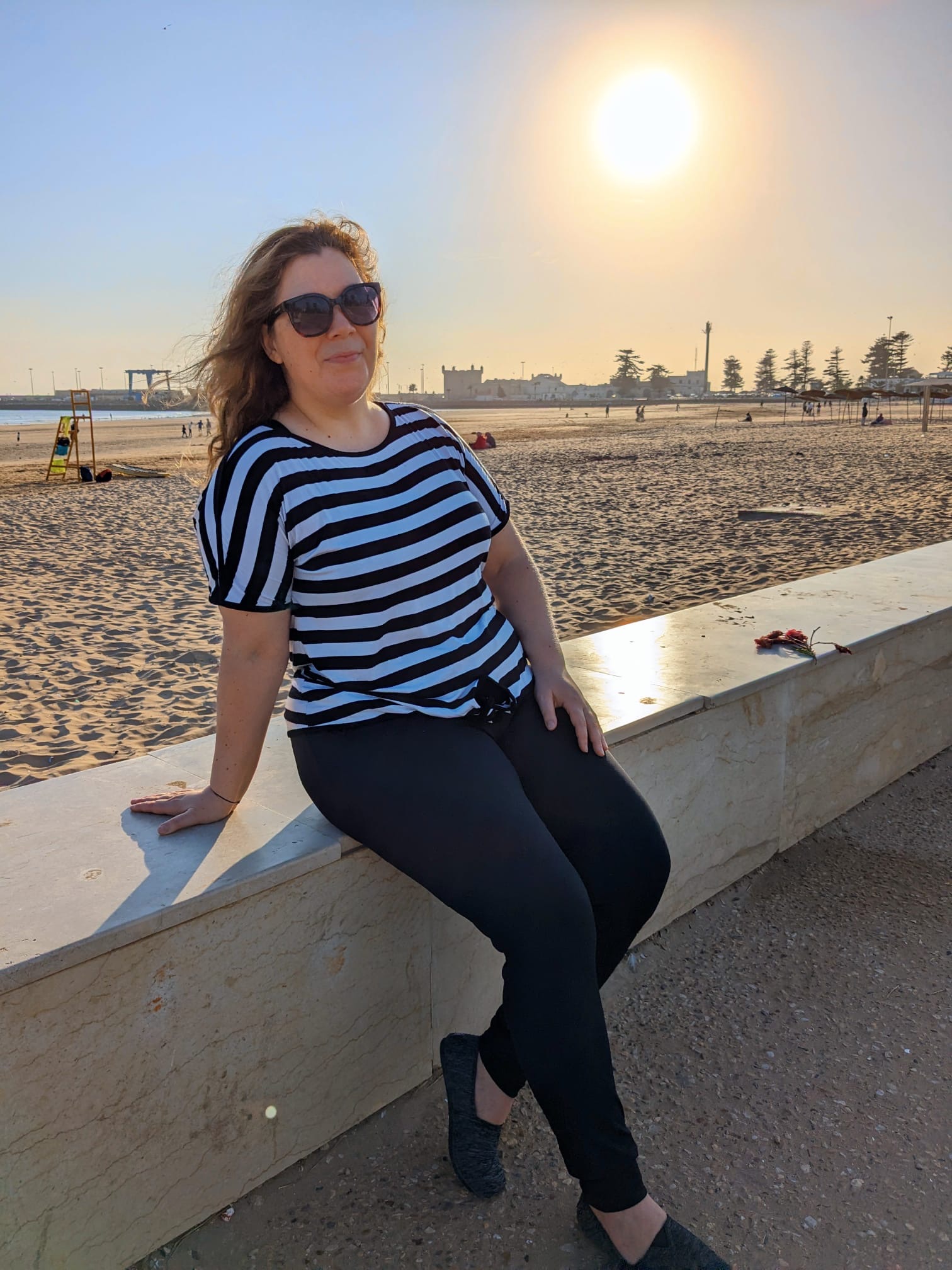 Woman sitting on a ledge with the beach and sun behind her